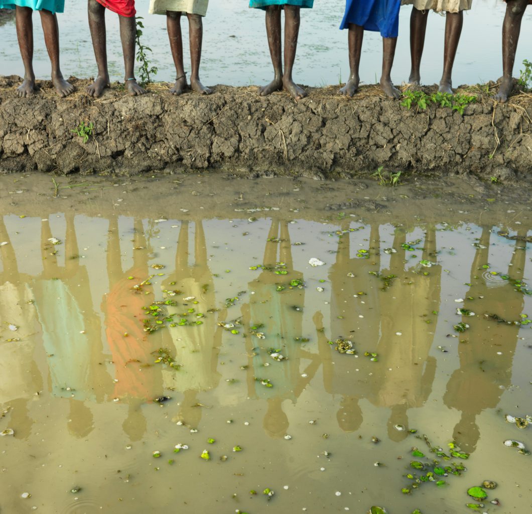 People stood in by flooded fields in South Sudan, only feet and water is showing, with community members reflections in the water