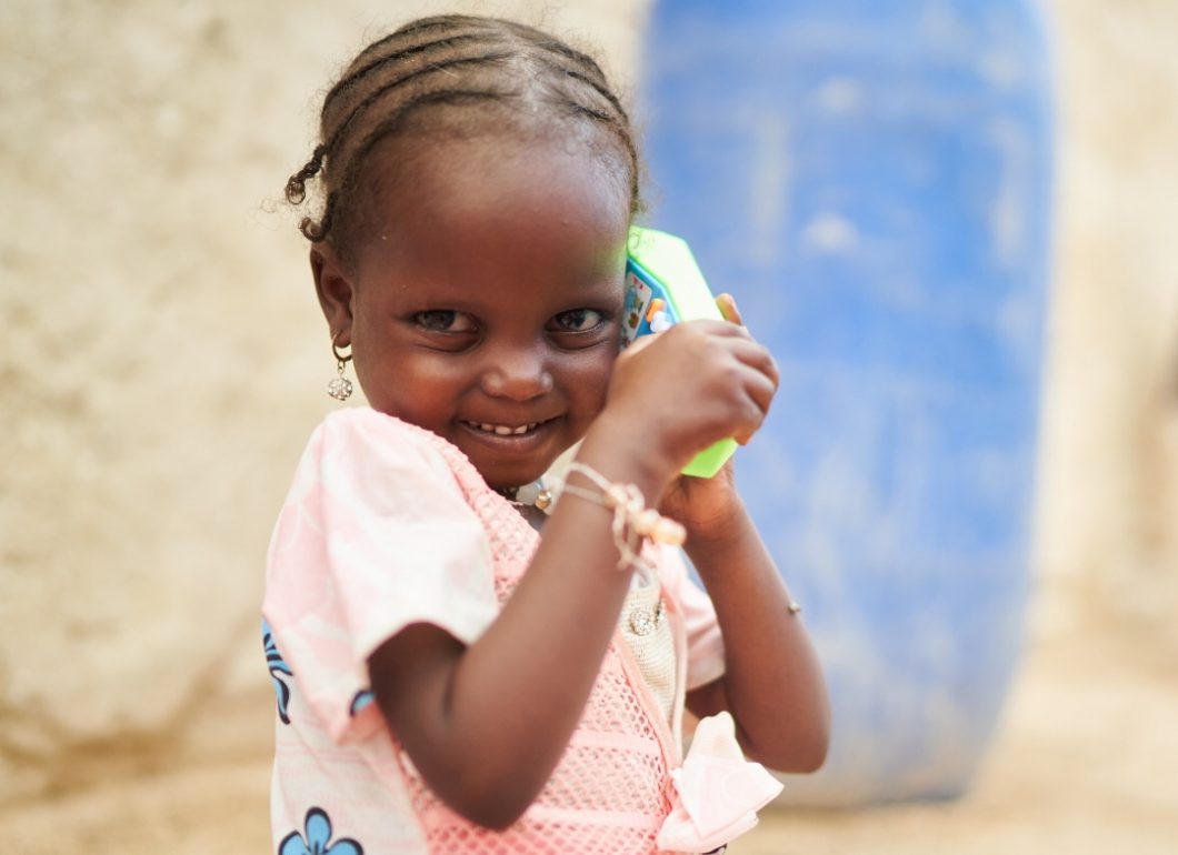 Achta, a young child in Chad, smiling brightly holding a green toy phone.