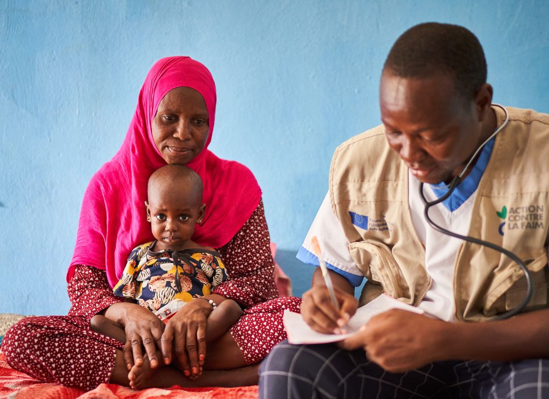 Koubra and her mother sit on a bed in Mao hospital. Dr. Patale, and Action Against Hunger doctor, is sat on the bed next to them writing notes.