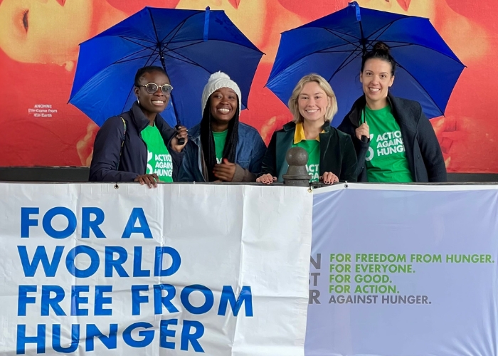 Four superstars in Action Against Hunger t-shirts behind banner under blue umbrellas.